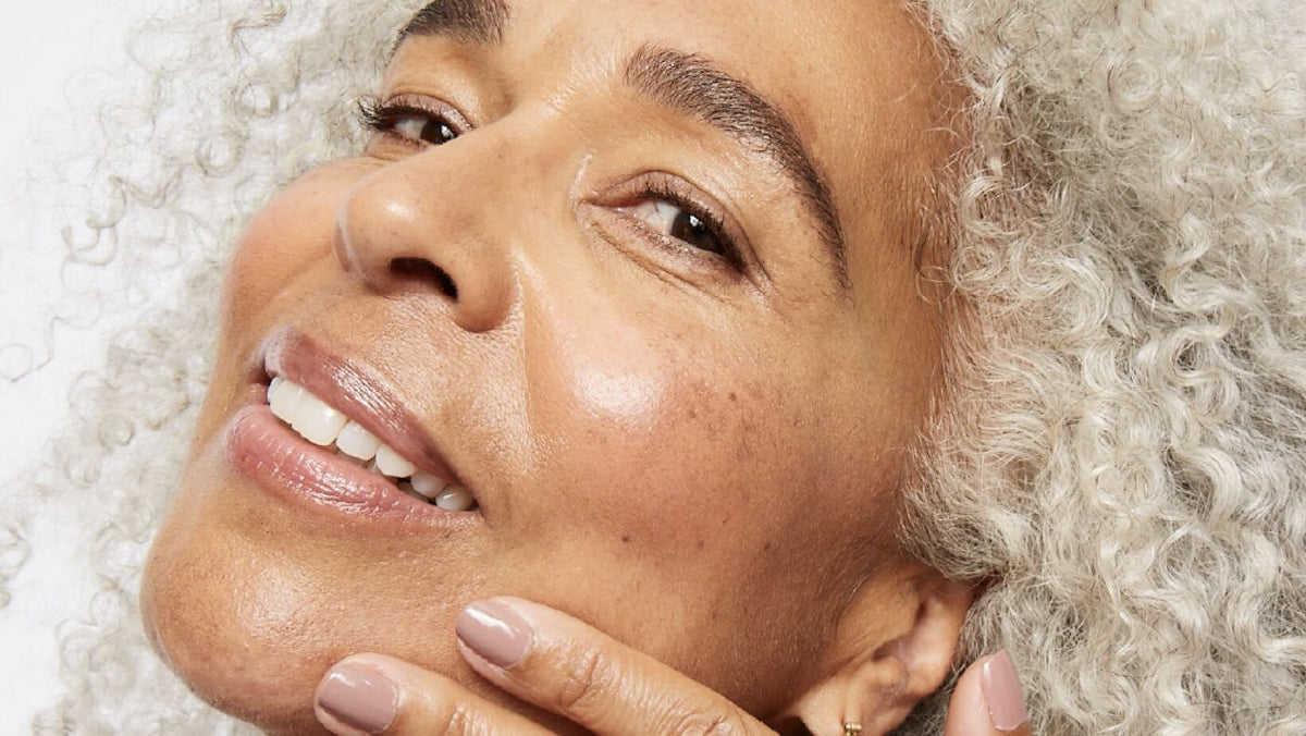 A beauty close-up highlighting glowing, natural-looking skin, a soft lip gloss, and full, textured gray curls against a bright white background.